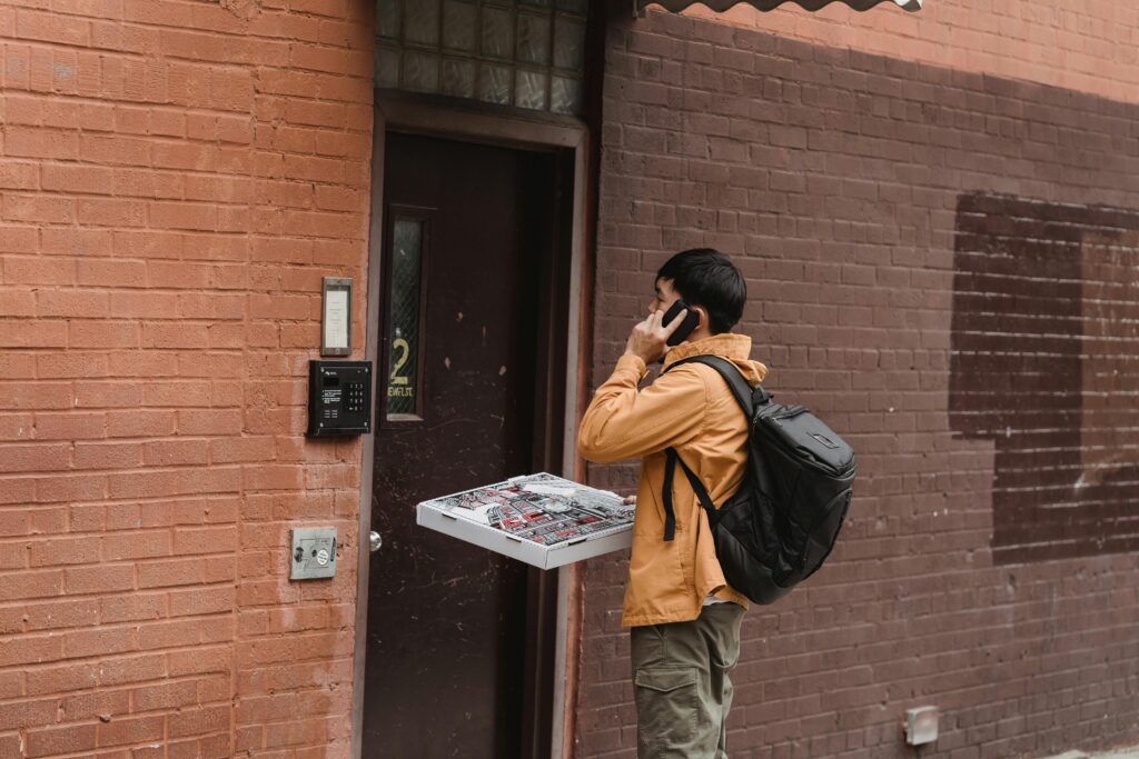 Man delivering pizzas, talking on cellphone, standing at apartment door with delivery bag.
